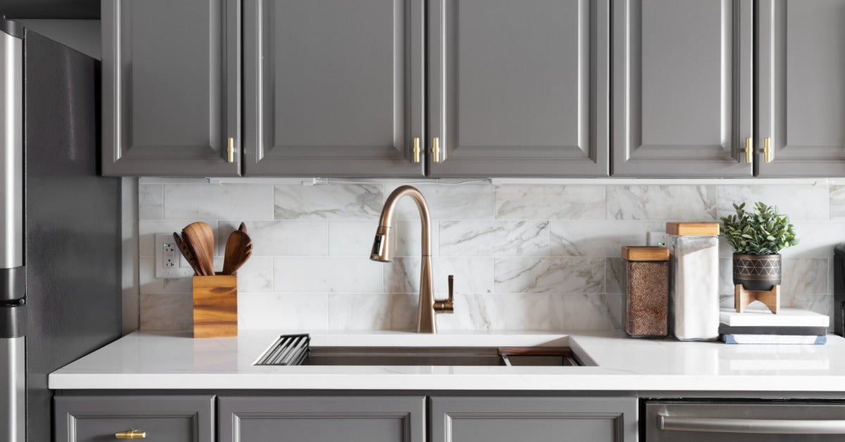A modern residential kitchen with gray cabinets and a sleek white countertop. The kitchen has a marble backsplash.