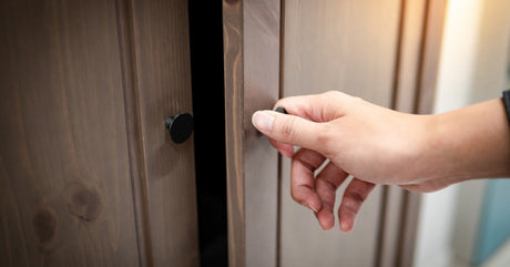 A person opening a brown wooden closet door in their home. The closet is installed in a brightly lit room.