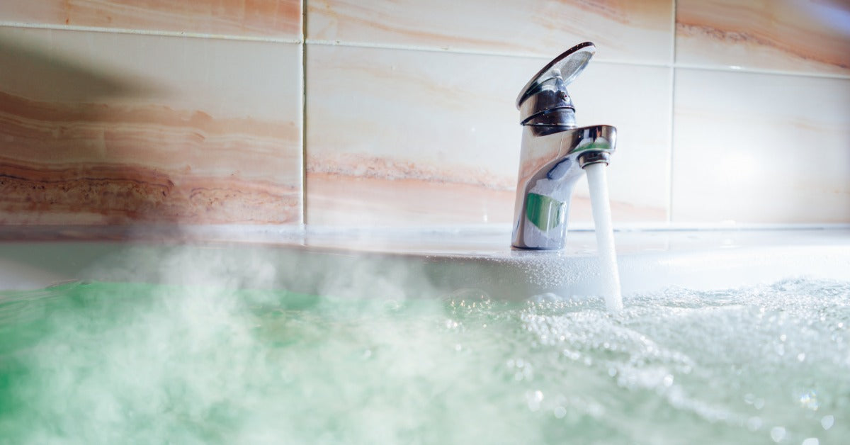 A sleek metal faucet filling a bathtub full of hot water. Steam rises from the blue water in the bathtub.