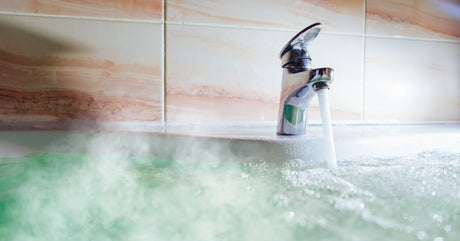 A sleek metal faucet filling a bathtub full of hot water. Steam rises from the blue water in the bathtub.