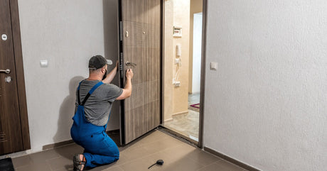 A contractor wearing blue overalls and a hat working on a large beige interior door inside a brightly lit home.