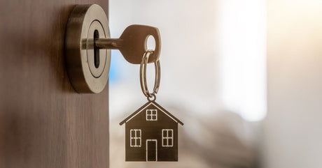 A metal key inside an open wooden door that leads into a home. The keychain is shaped like a small house.