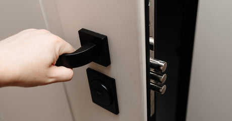 A person opening a gray door inside a home. The interior door has a long, black handle and a black lock.