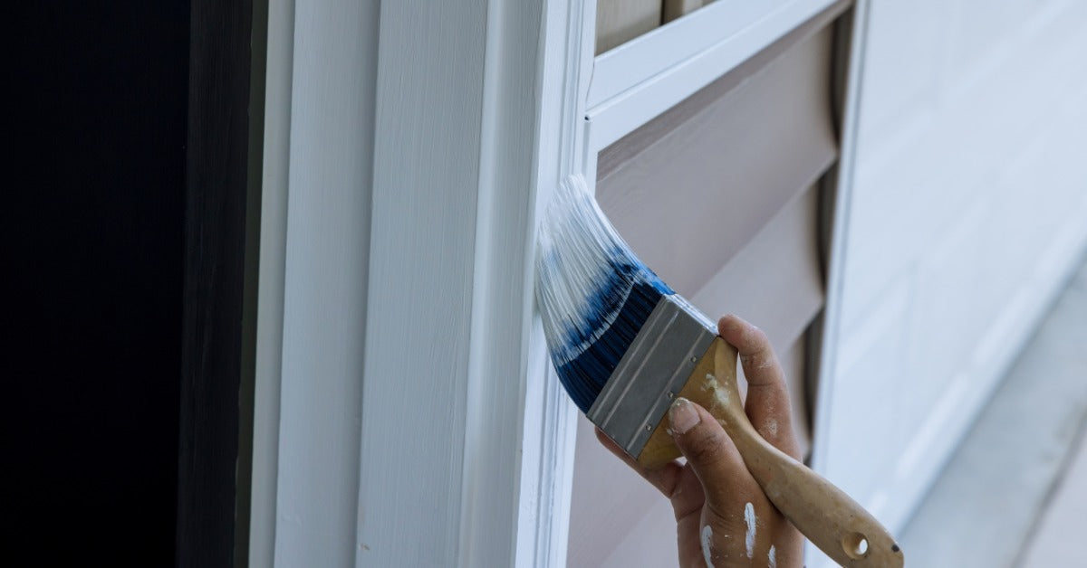 A person painting the outside of their wooden front door during the daytime. The paint on the brush is white.