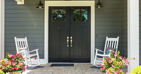 A front porch with a big black door, two white rocking chairs, and two potted plants. The plants have pink flowers.