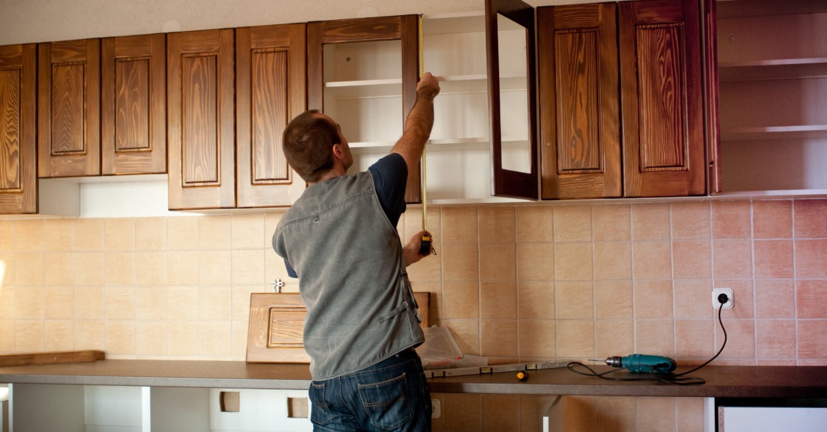 A carpenter holding measuring tape against a wooden kitchen cabinet inside a home. A drill lays on a nearby counter.