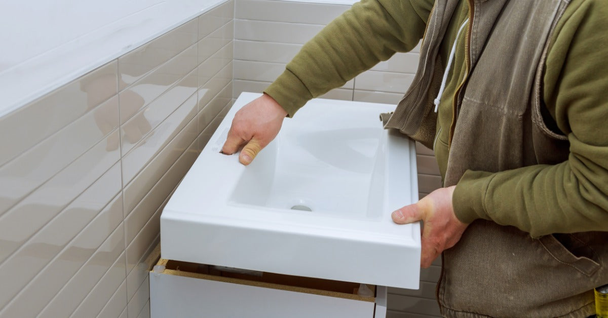 A contractor wearing a jacket and long-sleeve shirt installing a white bathroom vanity against a beige wall.