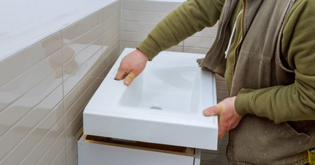 A contractor wearing a jacket and long-sleeve shirt installing a white bathroom vanity against a beige wall.