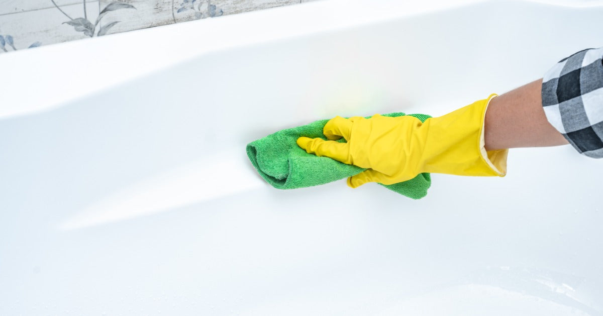A person wearing thick yellow gloves wiping a white acrylic bathtub with a large green microfiber cloth.